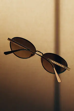 A pair of sunglasses with brown lenses and a golden frame rest on a beige surface, capturing a shadow of light and casting a shadow on the background.