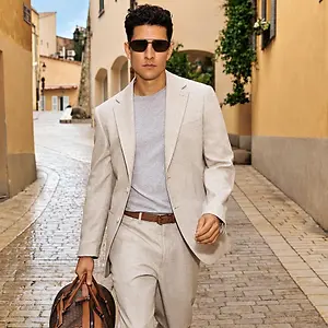 Man wearing sunglasses and a beige suit walking with a leather bag on a cobblestone street