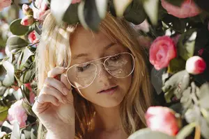 A blonde woman wearing clear glasses is standing in front of a cluster of pink flowers.