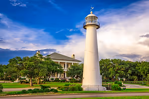 A lighthouse stands tall near a house with a gray roof on a grassy field under a partly cloudy sky