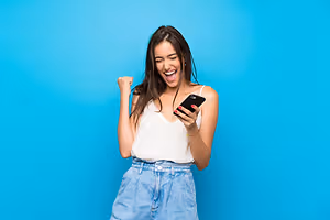 A woman smiling and celebrating while holding a phone and wearing a white sleeveless top and denim shorts against a blue wall