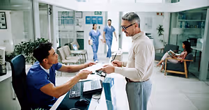 A man is talking to a man in blue scrubs in a hospital. The man in scrubs is sitting on a chair and holding a paper and a pen. The man is holding a paper and a pen and is looking at the man in scrubs. There are other people in the background, including a woman sitting on a chair and a man in blue scrubs walking.