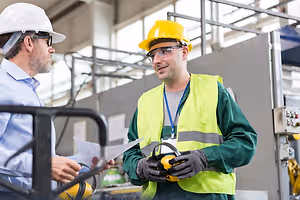 Two men in protective gear and hard hats stand in a factory setting, one holding a paper and the other holding a pair of ear protection