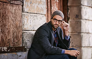 A man with a beanie and glasses is sitting on a brick wall and posing for a photo.