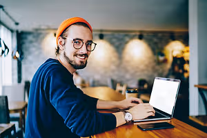 Man in blue long sleeve shirt and orange beanie sitting at a table in a cafe working on his laptop.