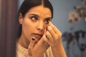 A woman wearing a white sweater and earrings is applying a contact lens in front of a mirror.