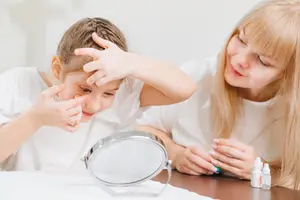 A young girl is looking into a mirror with a woman beside her, possibly applying makeup or skincare.
