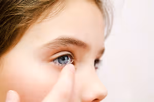 A girl with brown hair is trying to put on a contact lens in her right eye while looking at something in front of her.