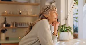 A woman wearing a sweater is sitting in her kitchen and looking out the window with her chin on her hand.
