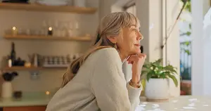 A woman wearing a sweater is sitting in her kitchen and looking out the window with her chin on her hand.