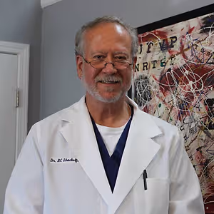 Dr. H.C. Shackelford, a medical professional, is standing in front of a white door and a wall with an abstract painting. He is wearing a white coat with the text 'Dr. H.C. Shackelford' on it, glasses, a necklace, and a blue shirt. He is smiling and seems to be posing for a photo.