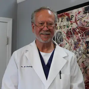 Dr. H.C. Shackelford, a medical professional, is standing in front of a white door and a wall with an abstract painting. He is wearing a white coat with the text 'Dr. H.C. Shackelford' on it, glasses, a necklace, and a blue shirt. He is smiling and seems to be posing for a photo.