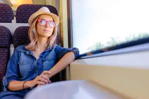 A woman is sitting in a train wearing a denim shirt and a hat with a bracelet on her left hand looking out the window