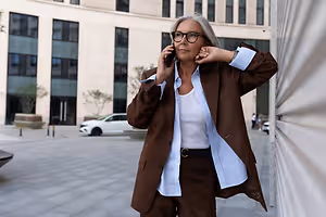 A woman wearing glasses and a brown suit stands on a city street, holding a cell phone to her ear while looking ahead.