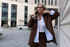 A woman wearing glasses and a brown suit stands on a city street, holding a cell phone to her ear while looking ahead.