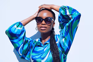 African woman posing for a photo wearing a blue and green patterned shirt, sunglasses, and earrings