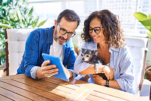 A man and a woman sitting on a chair in front of a table, looking at a tablet with a dog on the woman's lap.