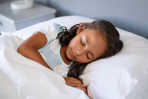 A young girl sleeps soundly on a bed covered with a white blanket, wearing a blue and white shirt, with a nightstand in the background.