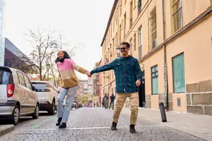 A man and woman are holding hands and laughing on a city street, with buildings and parked cars in the background.