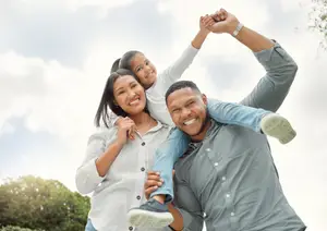 A man and woman are smiling and holding a young girl in a park with trees and a blue sky in the background