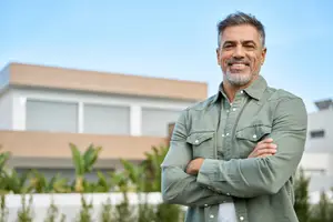 A man is smiling and standing in front of a house with his arms crossed.