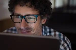 A man wearing glasses with a beard and mustache is looking at his laptop with a smile on his face.
