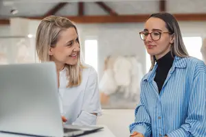 Two women looking at a laptop in an office setting, smiling and engaged in conversation.