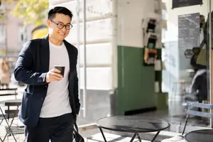 A man in a black blazer and white t-shirt walks in a coffee shop and holds a coffee cup.