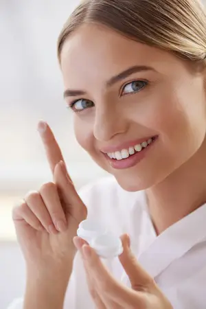 A woman in a white lab coat is holding a contact lens and pointing at her eye with her finger