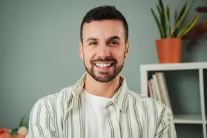 A smiling man with a beard and mustache wearing a striped shirt poses for a photo against a light green wall with a plant and books in the background