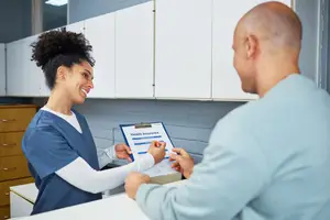 A woman in a blue scrub uniform is smiling and looking at a man in a blue shirt as she hands him a clipboard with the words Health Insurance written on it.