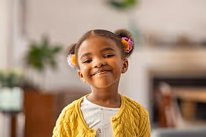 A young girl with a yellow cardigan and pink and yellow flower clips in her hair smiles in a living room