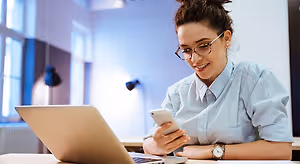A woman wearing glasses and a blue shirt is smiling and looking at her phone while sitting in front of a laptop on a desk.