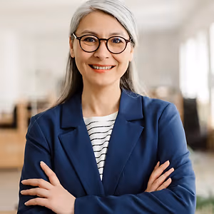 A woman with gray hair wearing glasses and a blue blazer is smiling and posing for a photo in an office.