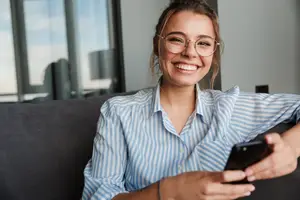 A woman in a striped shirt and glasses is smiling and using her phone on a couch