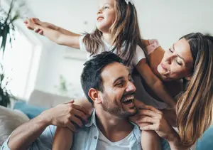 A family is laughing on a couch with a young girl on her father's shoulders.