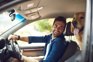 A man is driving a car with his dog sitting on the passenger seat and his tongue out