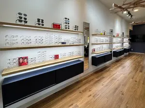 A clean and modern eyeglasses shop with wooden shelves, wooden floor, and hanging lamps. There are a variety of eyeglasses displayed on the shelves, including sunglasses and prescription glasses. A large mirror is mounted on the wall, and there is a picture of a couple on the shelf. The shop has a modern and elegant design, with a focus on showcasing the eyeglasses.
