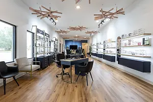 A well-lit indoor space resembling an optometrist's office, featuring modern furniture, a wooden floor, and a white wall with shelves displaying eyeglasses and other items.