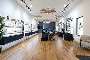 The interior of a modern optometrist office with wooden floors and white walls. There are shelves with sunglasses, a desk with a computer, a chair, and a couch. There are also lamps hanging from the ceiling and a potted plant on the left. The room has a glass window on the right, and there is a view of the outside.