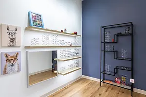 A room with a white wall, wooden floor, and blue wall with shelves, a mirror, and a framed picture of a fox wearing glasses. The shelves have eyeglasses, and the black shelf has a display of glasses and other items.