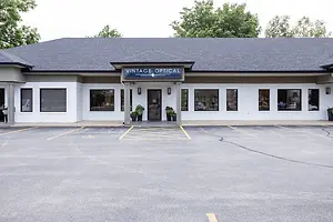 Vintage Optical building with a gray roof and white walls, featuring a sign, potted plants, and glass windows, with an empty parking lot in front under a clear sky.