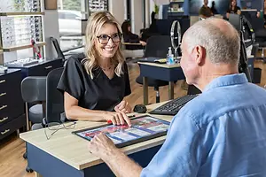 A woman in glasses sits at a desk with a tablet while talking to a man in a blue shirt in an office with other workers in the background