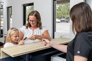 An adult woman and a young girl standing in front of a counter, holding a credit card and smiling at a staff member who is also smiling at them. There are chairs, a printer, and a glass door behind them, and a car parked outside.