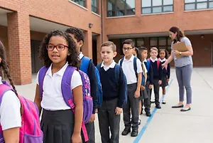 A group of children standing in a line in front of a building with a woman holding a folder