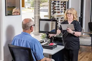 An adult male wearing glasses is sitting on a chair and looking at a monitor while an adult woman wearing glasses and a bracelet is standing in front of him and holding a tablet and a monitor.