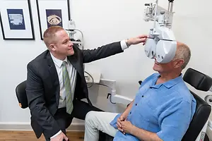 A man with glasses is adjusting an older man's eyeglass frames using an ophthalmoscope in an office setting.