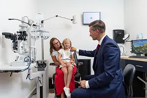 A young girl in a white dress is sitting on a chair while a man in a blue suit is adjusting her glasses in an eye clinic room.