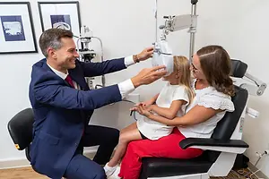 A man in a blue suit is adjusting an eye exam machine on a young girl sitting in a chair with her mother who is wearing glasses and a white blouse.