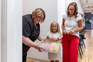 Three women smiling while holding a basket of toys in a hallway.